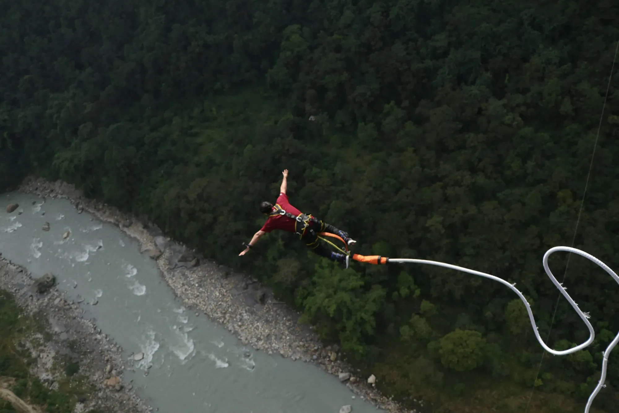 A bungee jumper in a harness diving above a grey-green river in a steep forested gorge