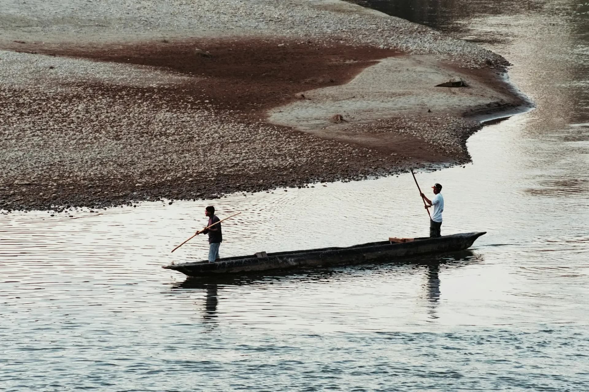 Two boatmen poling a dugout canoe past a gravel bar on a wide river