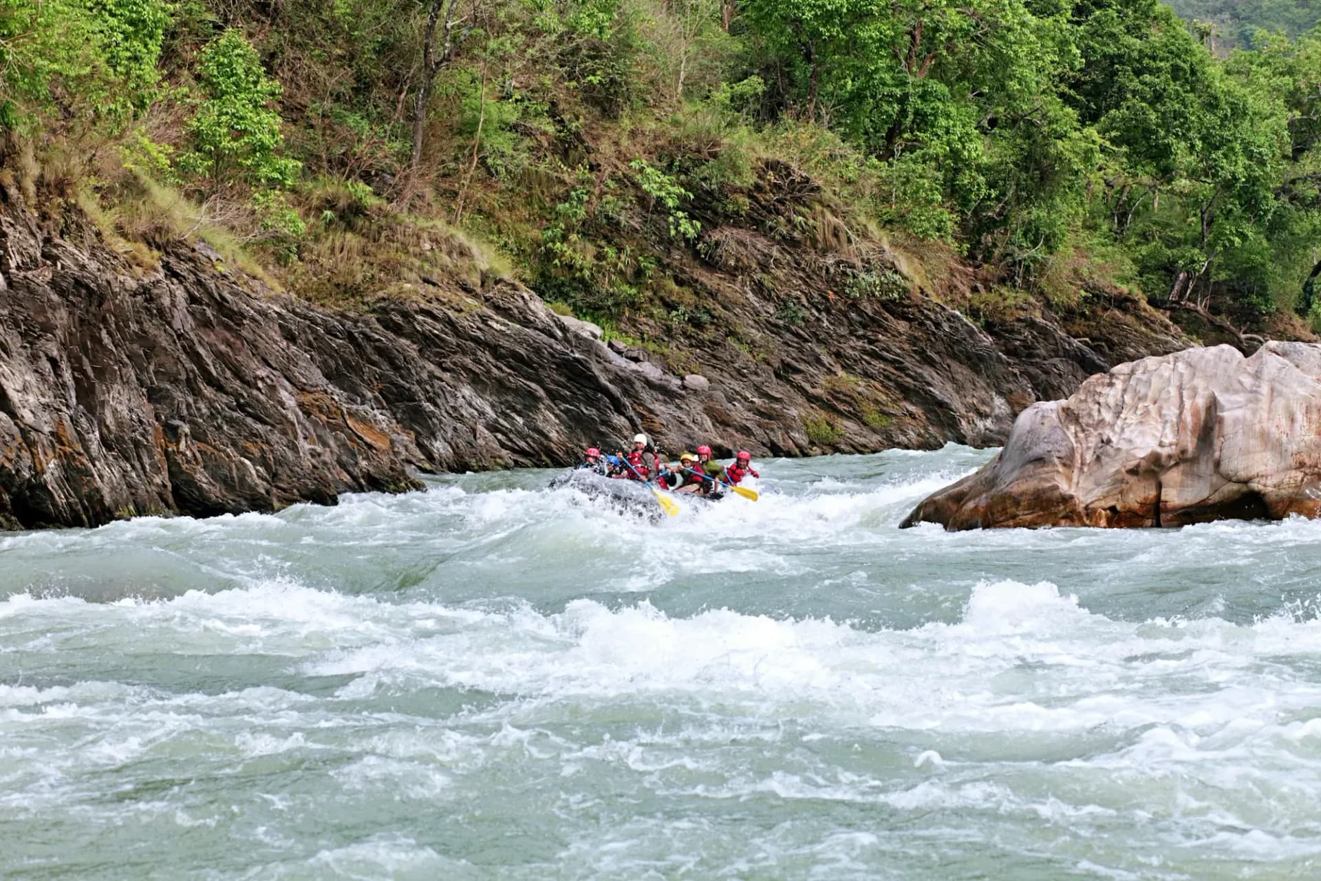 A raft of paddlers in white water between a layered rock wall and a large boulder