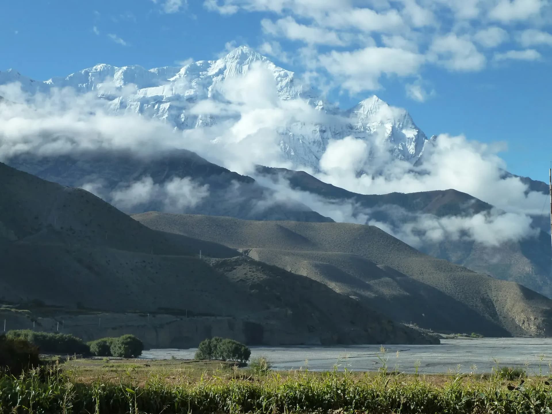 Snow peaks rising through cloud above a wide riverbed and green fields