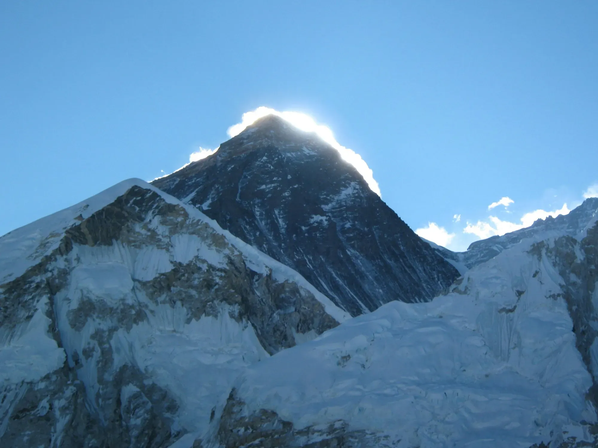 The dark rock summit pyramid of Everest above snowy ridges, with the sun glowing behind it