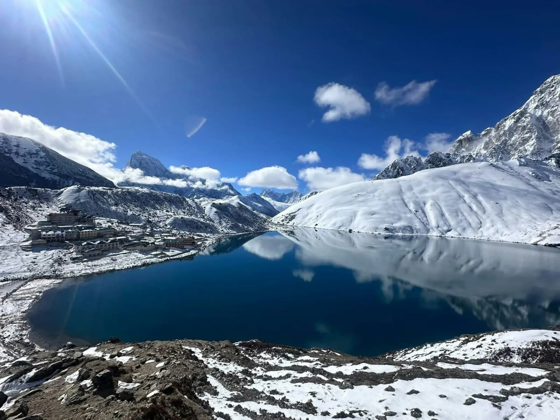 A still, deep-blue Gokyo lake reflecting snowy hills, with lodges on the shore
