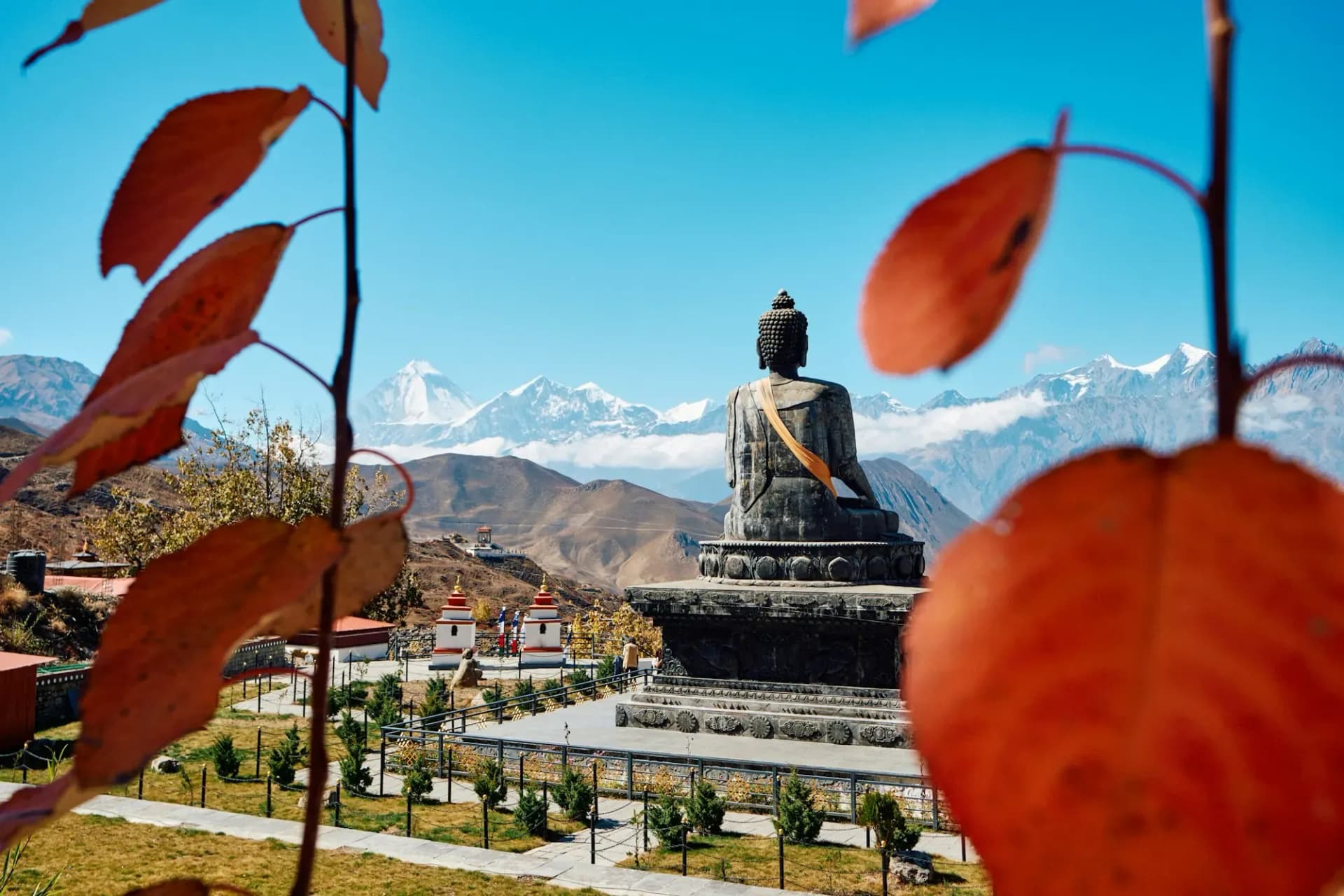 Large Buddha statue facing snow peaks near Muktinath, framed by autumn leaves