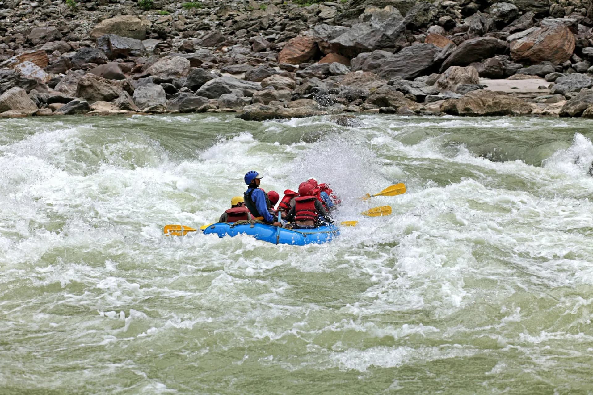 A blue raft with a helmeted crew pushing into a white-water wave below boulders